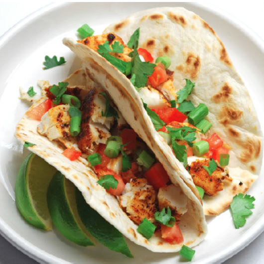 Two flour tortilla tacos filled with tilapia, tomatoes and cilantro on a plate garnished with lime wedges.